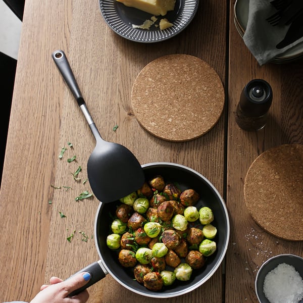 Es ist ein Tisch mit einer KAVALKAD Bratpfanne in Schwarz zu sehen. Darin befinden sich Rosenkohl und HUVUDROLL Proteinbällchen. Die Pfanne steht auf einem HEAT Topfuntersetzer aus Kork.