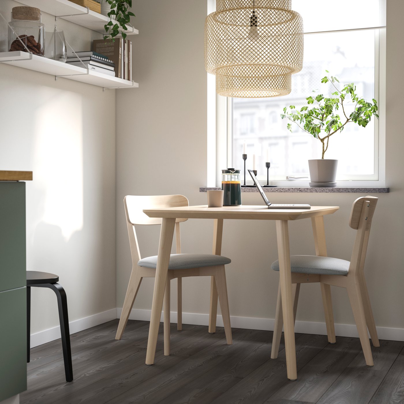 Kleiner, heller Essplatz mit rundem LISABO Tisch aus hellem Holz und zwei passenden Stühlen mit grauen Sitzpolstern, darüber eine große geflochtene Pendelleuchte, am Fensterbrett eine grüne Pflanze und auf dem Tisch eine Kaffeekanne, Tasse und aufgeklappter Laptop.