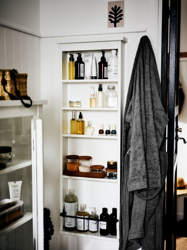 The corner of a white bathroom with a HEMNES wall shelf on one wall. Toiletries and bathroom accessories are on the shelves.