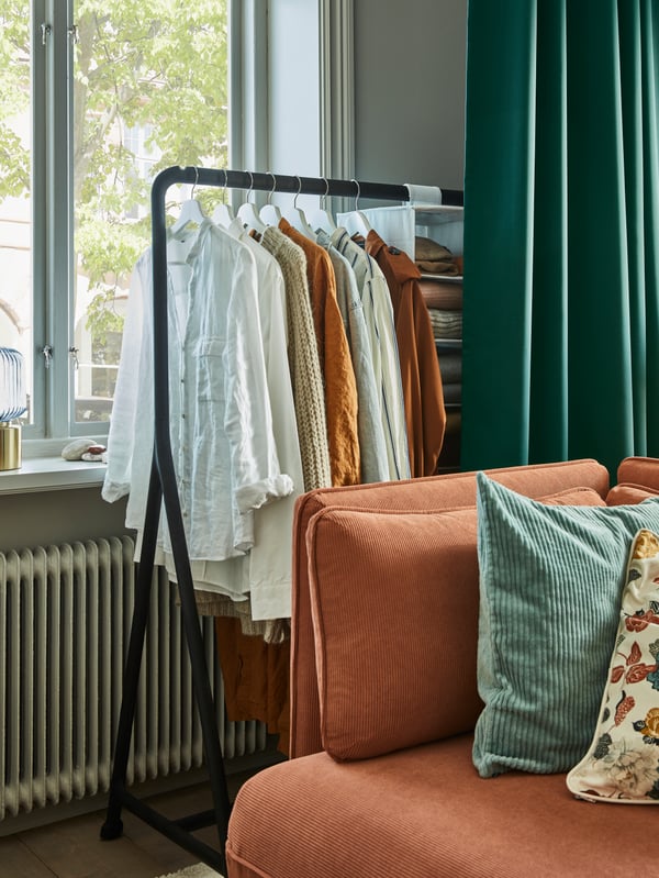A black standing clothing rack with hanging clothing is positioned against a large window next to a red sofa.