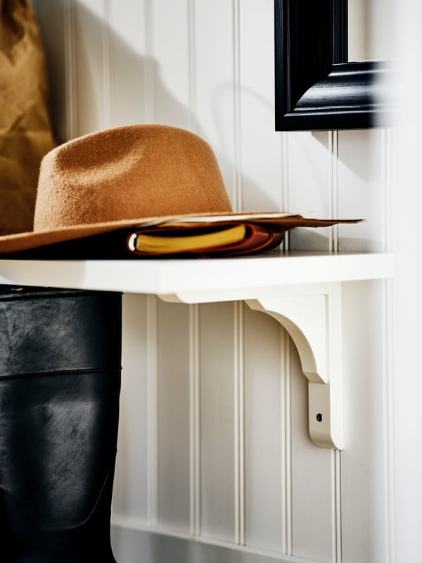 The end of a white BERGSHULT/RAMSHULT wall shelf on a wood-panelled wall with a brown hat and a notebook on it.