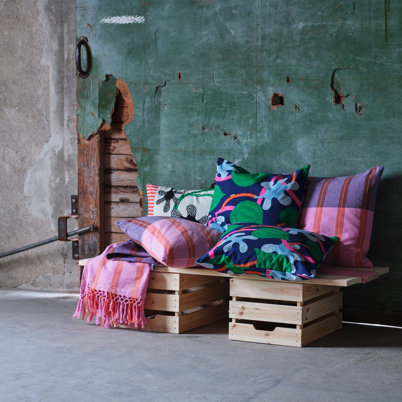 On top of a couple of wooden boxes are cushion covers and throws in pink, blue and green from the MÄVINN collection.