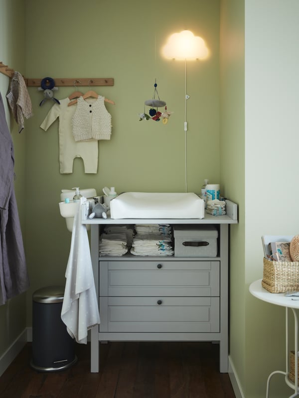 A grey changing table/chest of drawers, oak racks with baby clothes hanging, a babycare mat, a cloud white LED wall lamp.