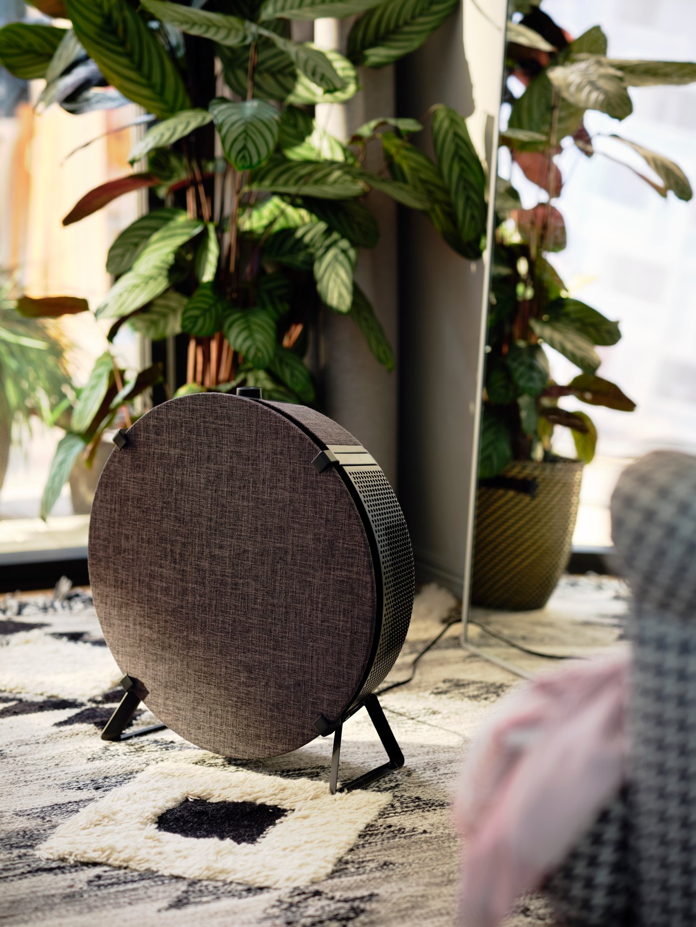 Ein STARKVIND Luftreiniger in Schwarz steht auf einem Teppich vor einer Pflanze. Der Luftreiniger lässt sich mit IKEA Home smart nutzen.