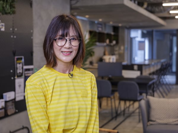 黄色のストライプシャツを着た人物が、モダンなオフィス風の室内に立つ。背景にテーブルと装飾