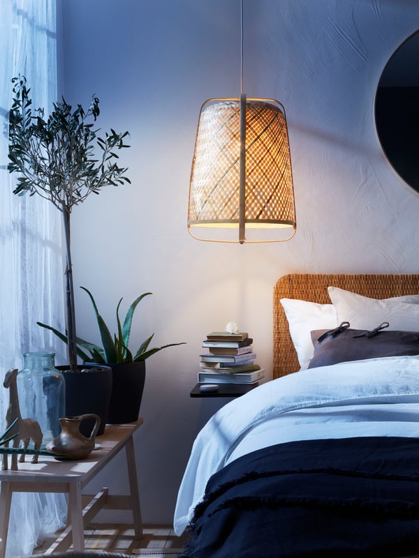 A hanging lamp with lamp shade above a bed, with plants by the window and books on a bedside table.