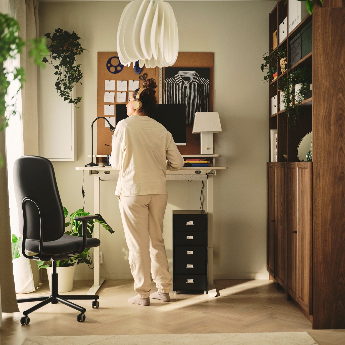 A home office with a beige TRAVSPORT desk sit/stand in oak effect, next to a black rolling chair and potted plants.