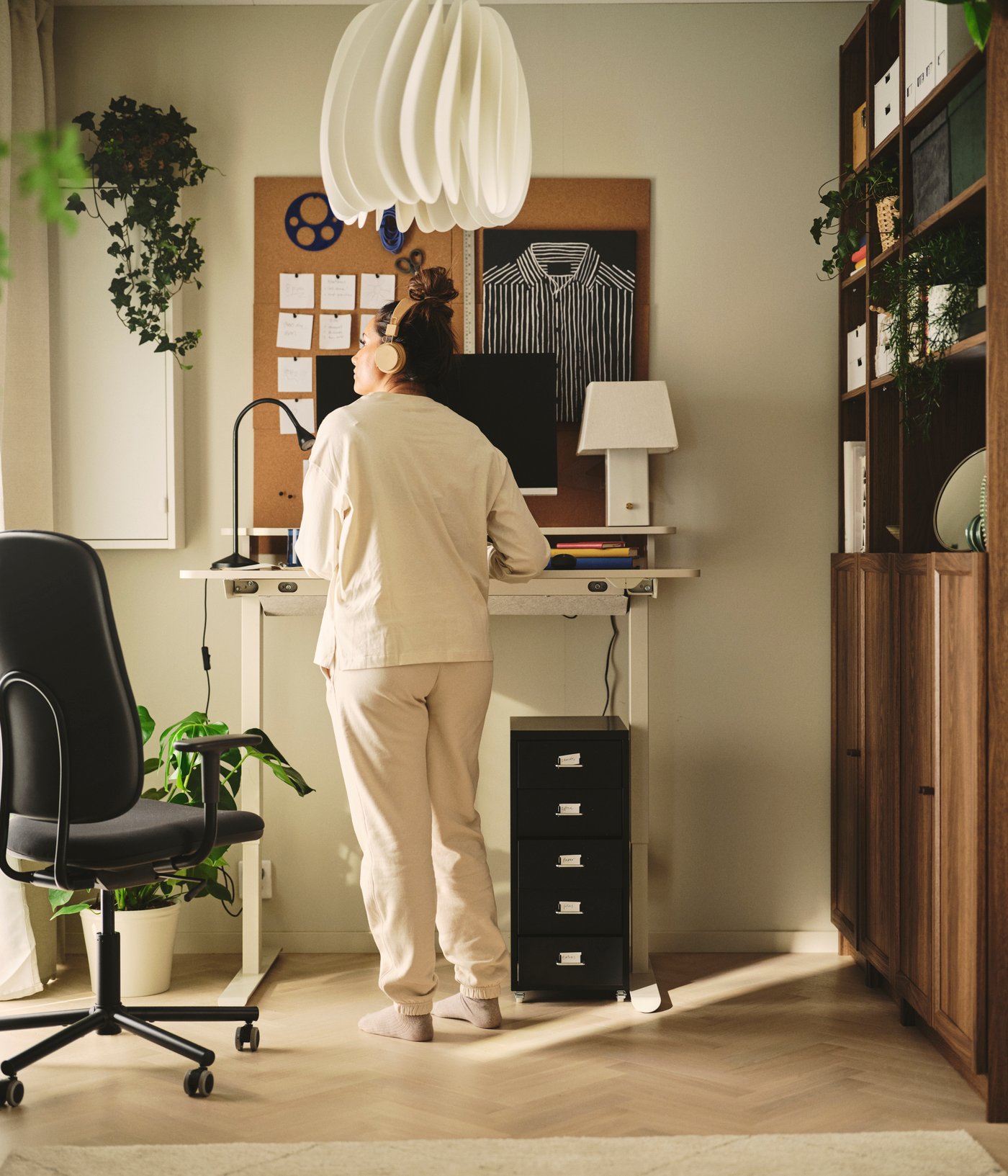 A home office with a beige TRAVSPORT desk sit/stand in oak effect, next to a black rolling chair and potted plants.