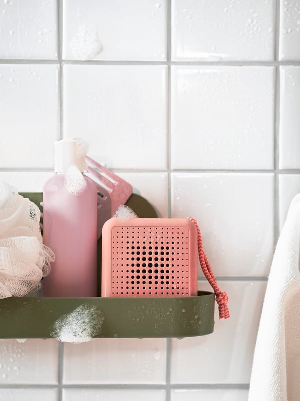 En ljusrosa VAPPEBY bluetooth-högtalare på en våt och tvålig duschhylla i ett vitkaklat badrum.
