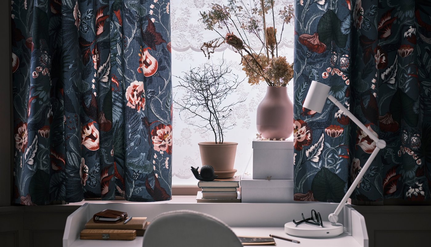 La ventana de una habitación con cortinas floreadas y en frente un escritorio pequeño con una lámpara blanca y otros artículos.