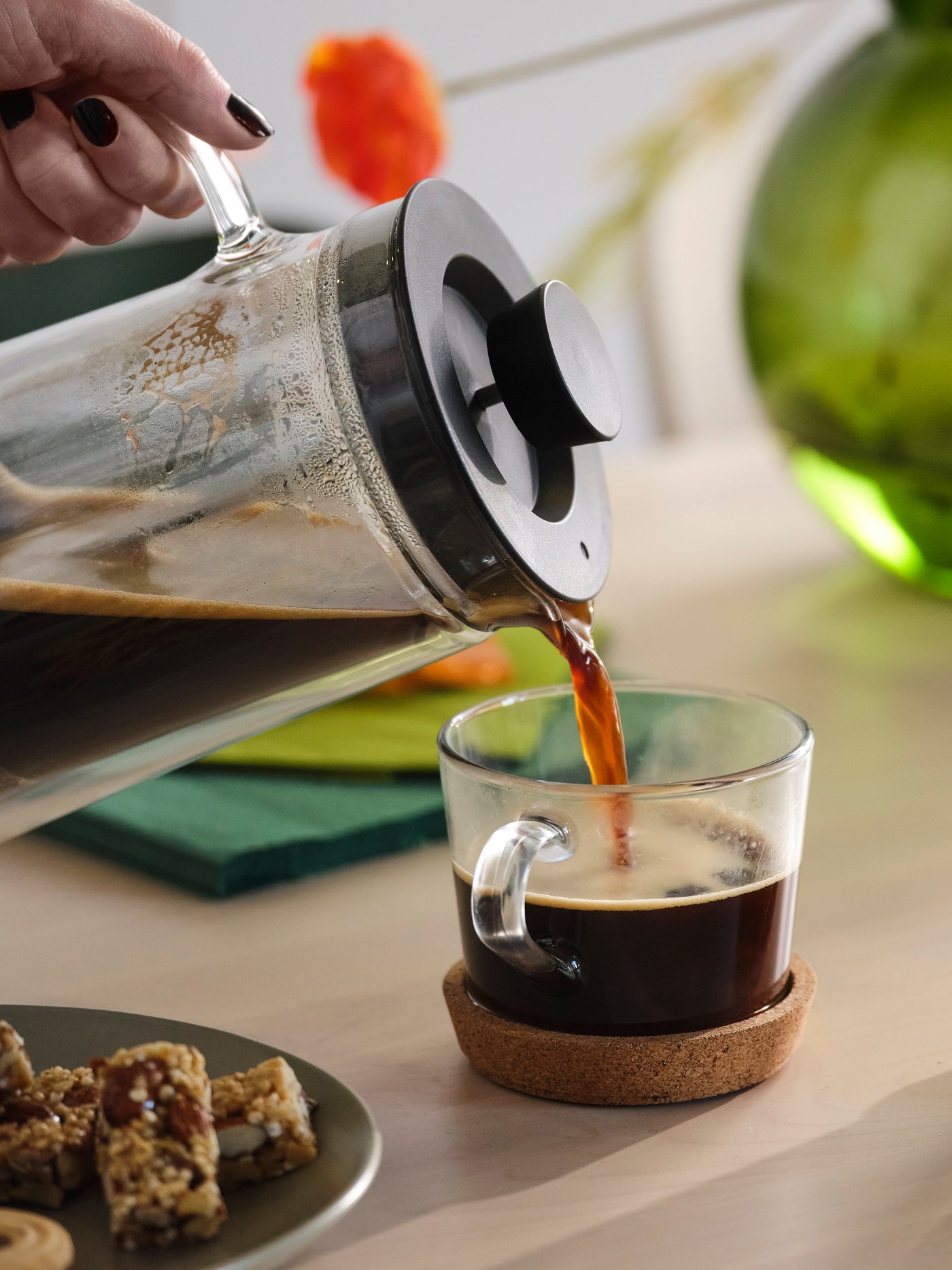 Kafa se sipa iz posude za kafu u šoljicu na podmetaču od plute. U pozadini su zelena vaza i narandžasti cvet.