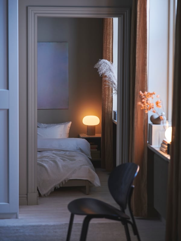 A black stained oak veneer FRÖSET easy chair stands in front of a doorway into a bedroom with a bed and a DEJSA table lamp.