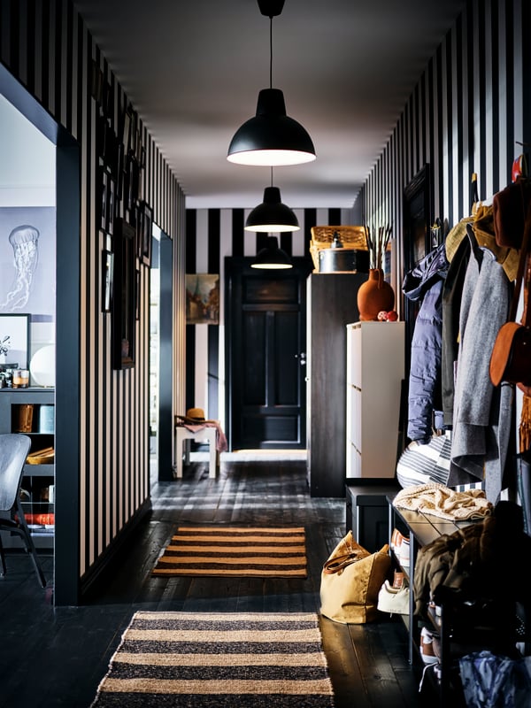 Un couloir avec des murs aux larges rayures en noir et blanc, avec des suspensions, des bancs range-chaussures PINNIG noirs et des tapis UGILT.