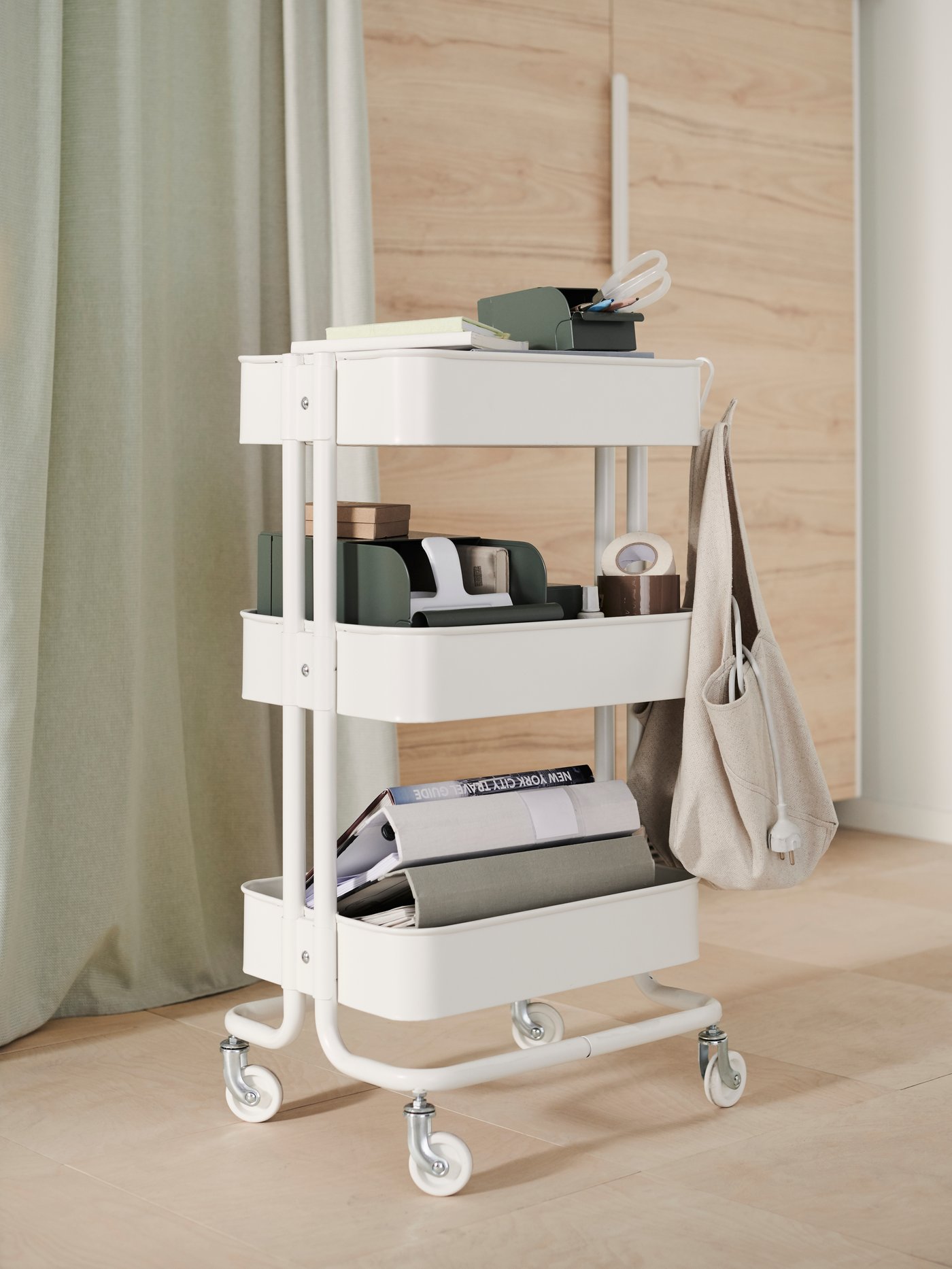 A white RÅSKOG trolley with four wheels filled with office supplies. A hook holds a natural NEREBY bag with computer cables.