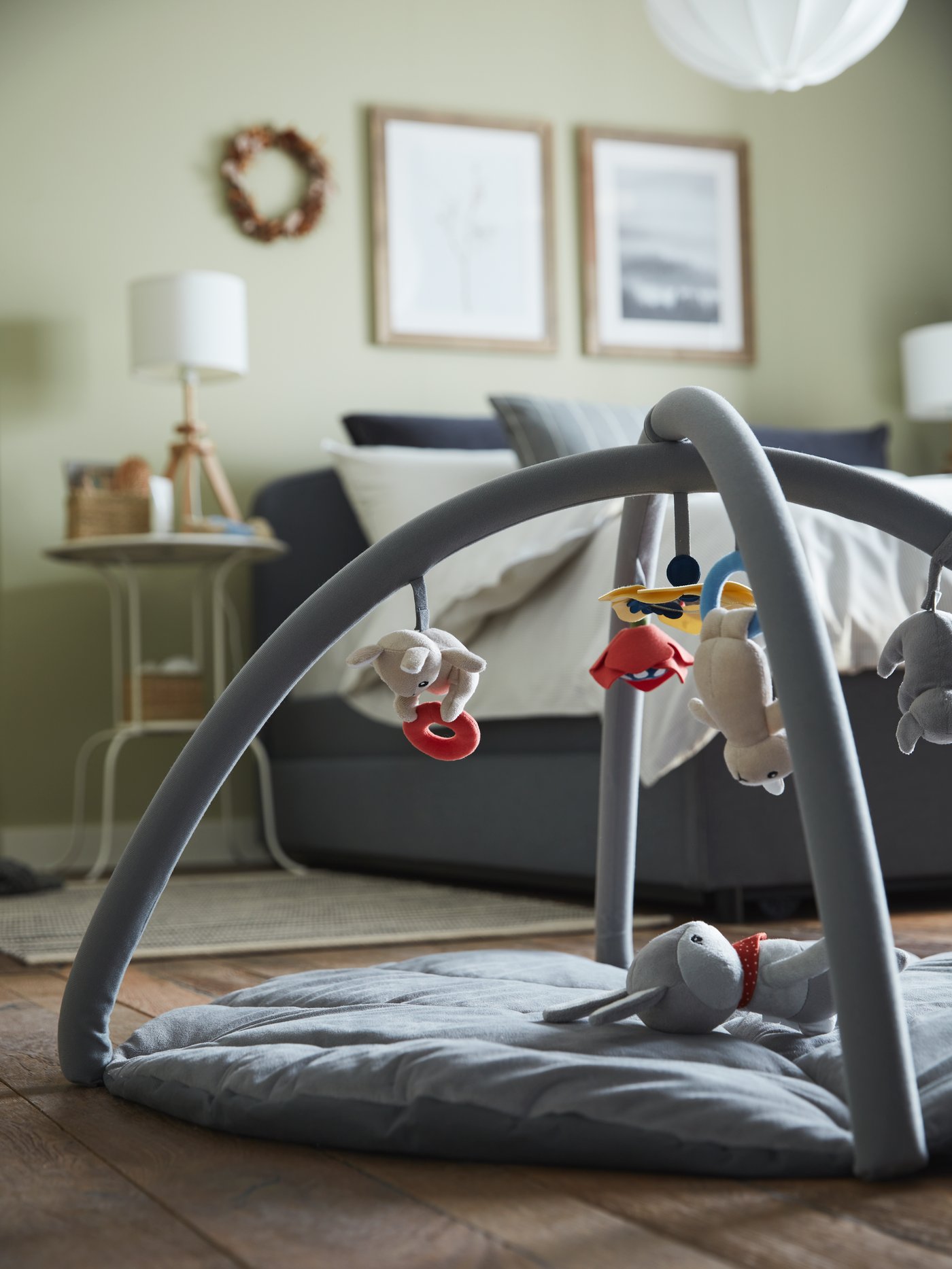 A multicolour baby gym on the floor, a white side table, a grey upholstered bed, an ash/white table lamp.