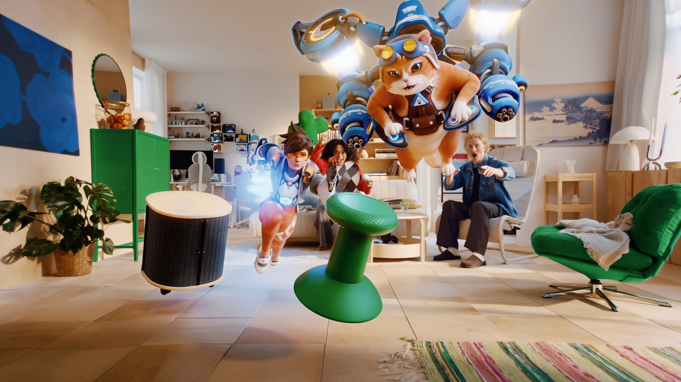 Overwatchi tegelased Tracer ja Jetpack Cat sööstavad läbi valgusküllase elutoa, samal ajal kui taustal on kaks elavat mängurit ning roheline analoogkangi meenutav taburet keerleb põranda kohal.				