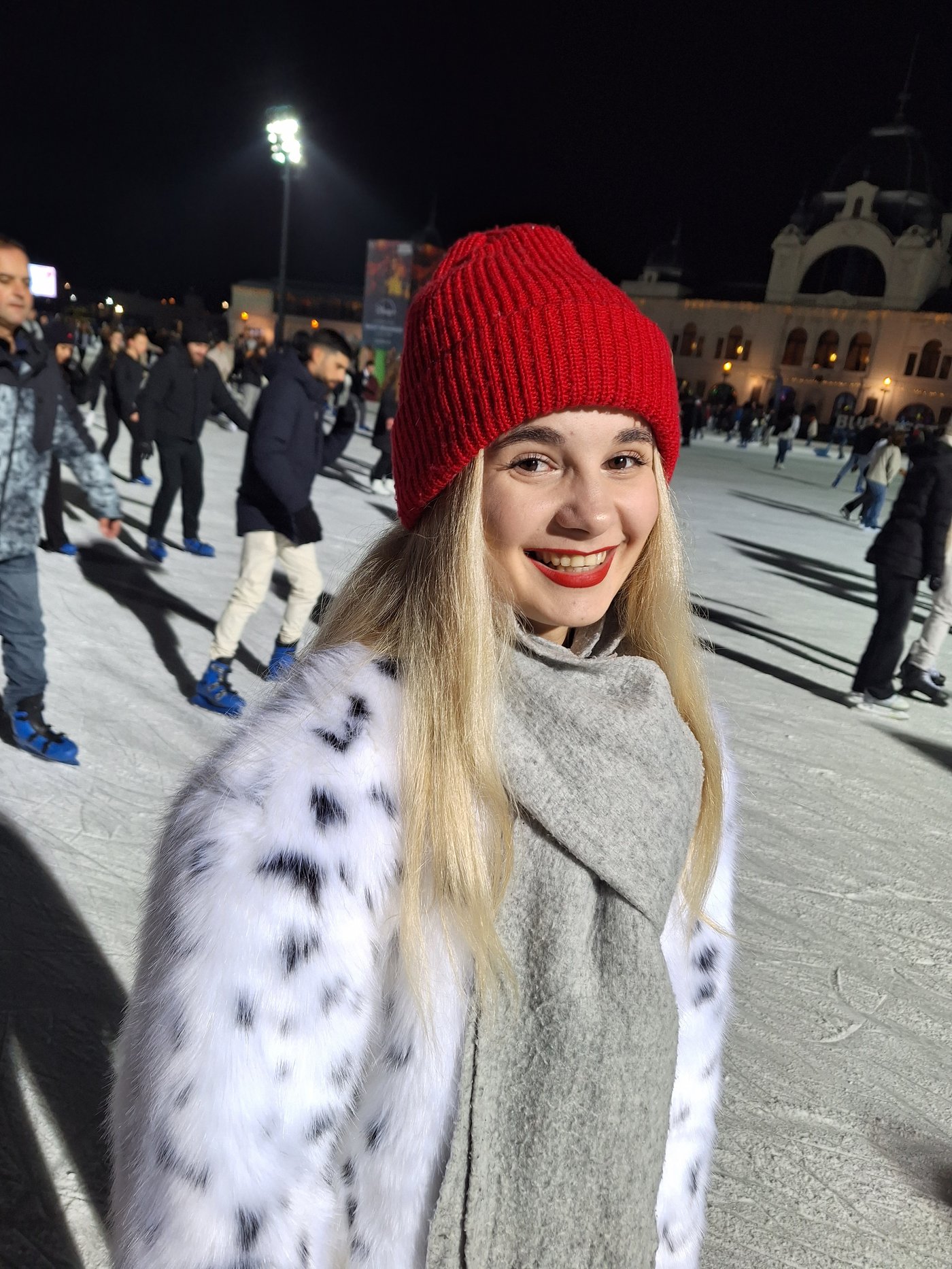 Yana na klizalištu s crvenom pletenom kapom i ljudi koji se klizaju u pozadini.