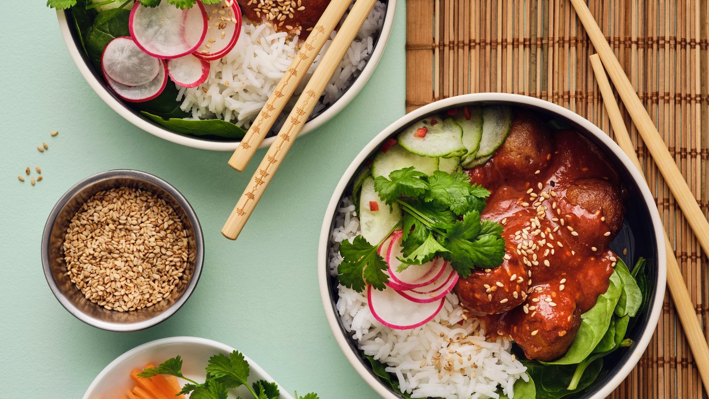 Two white bowls are placed on a light green table, and they are filled with a salad made of meatballs, rice and vegetables.