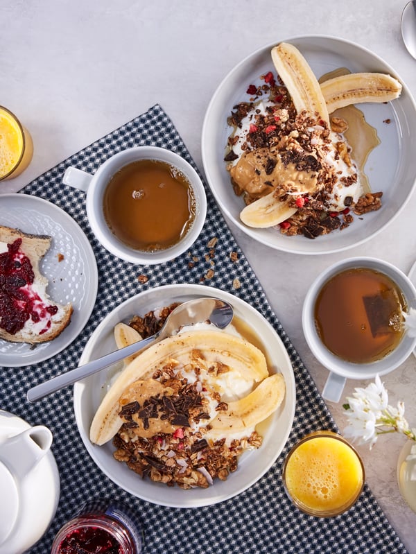 A breakfast table with two cups of tea, juice and two bowls of HJÄLTEROLL muesli with cocoa, dried berries and bananas.