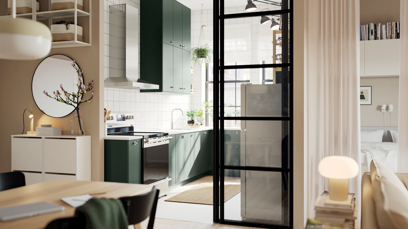 An open kitchen with deep green HAVSTORP kitchen fronts and wall cabinets up to the ceiling, next to a bright living room.