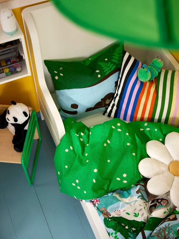 Cama infantil vista desde arriba con funda deplumón y almohada SKOGSDUVA, además de cojines y peluches alrededor.