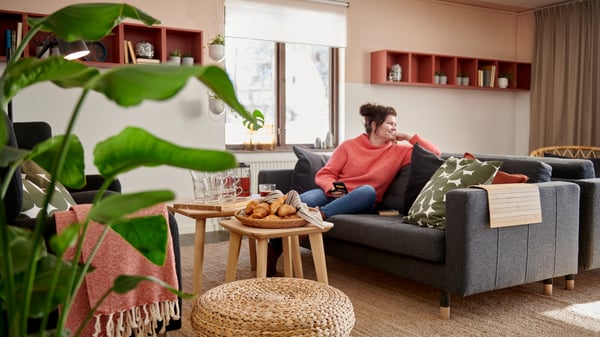 Mysig inomhusmiljö med person i en grå soffa med en mobil i handen, frukost på bordet och stora fönster i bakgrunden.