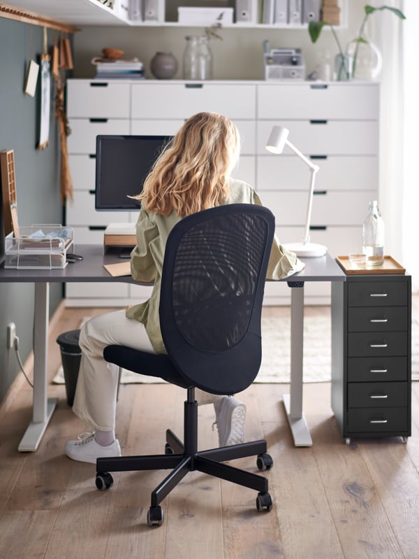 A person is sitting on a black FLINTAN desk chair at a desk with a white NYMÅNE desk lamp on it.