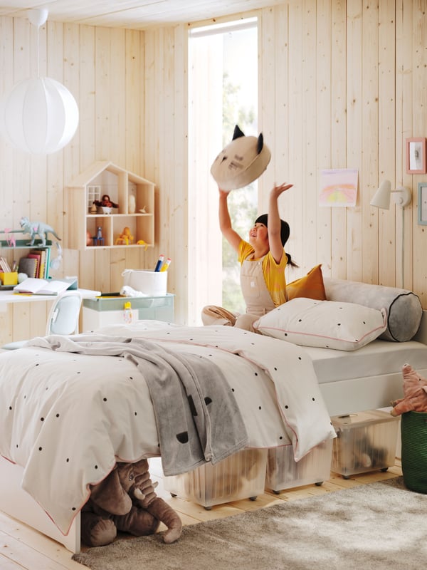 Young girl on bed with spotty bedding and storage under, playing with toy in bright children’s room with light wood walls.