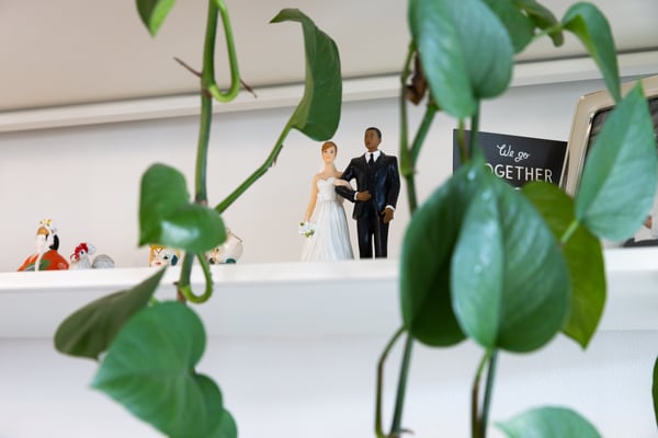 A view of some neat shelving, with a wedding cake topper and some other trinkets. Frames in the foreground of the photo is a green plant.