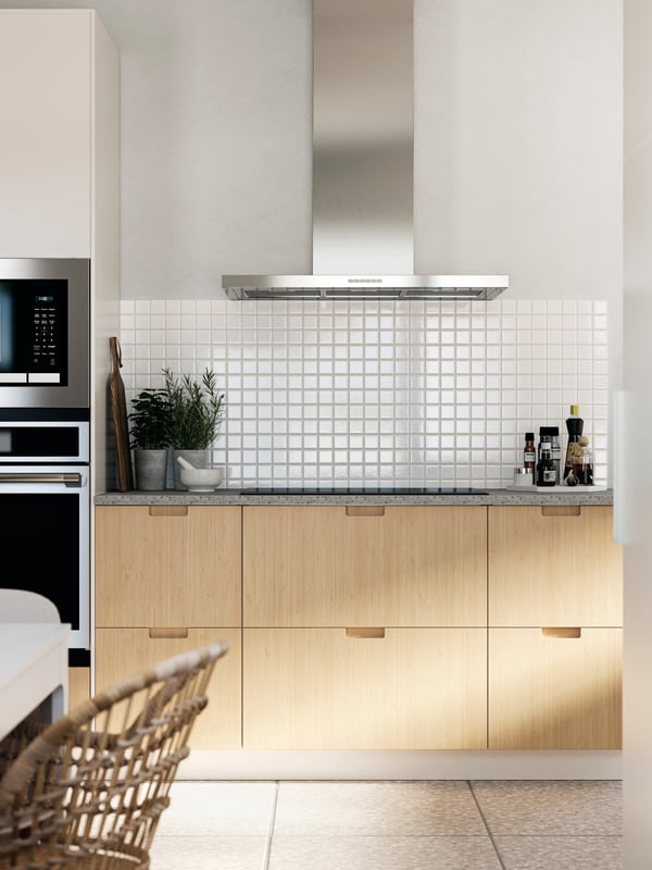 A kitchen with white walls, small white tiles, FRÖJERED kitchen fronts made from bamboo and stainless steel appliances.
