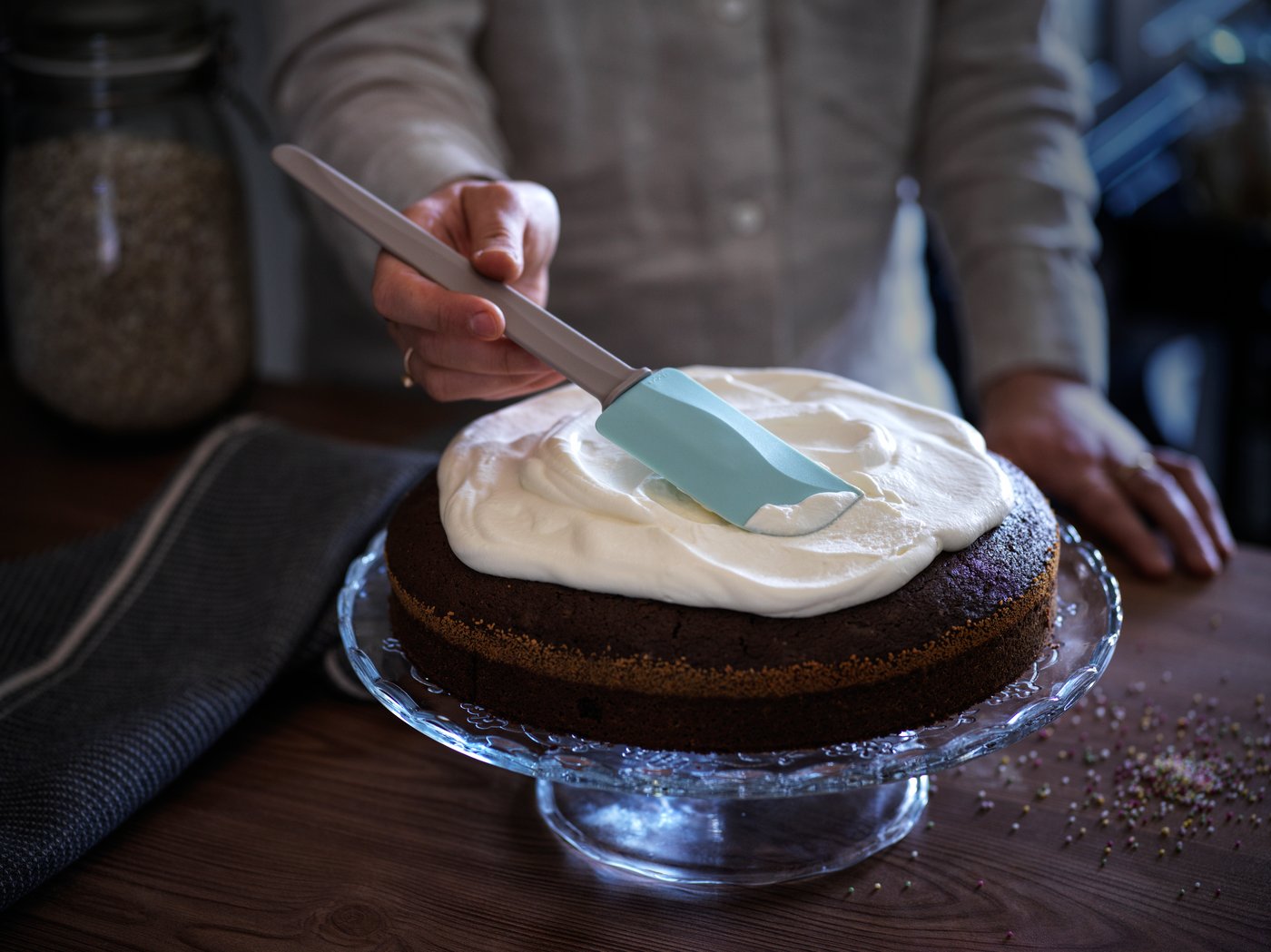 Baker uses rubber spatula to frost a cake on glass cake stand