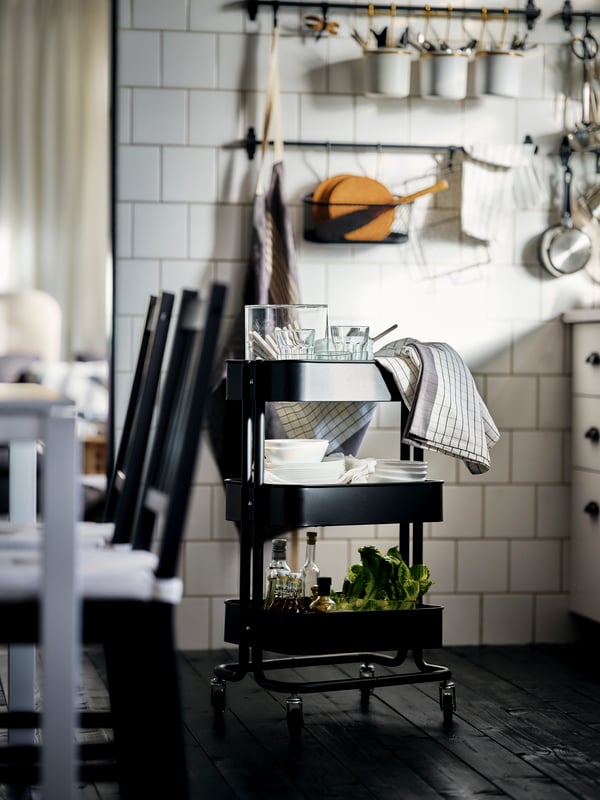 A black RÅSKOG cart holding dining accessories standing on a dark wood floor in a white-tiled kitchen.