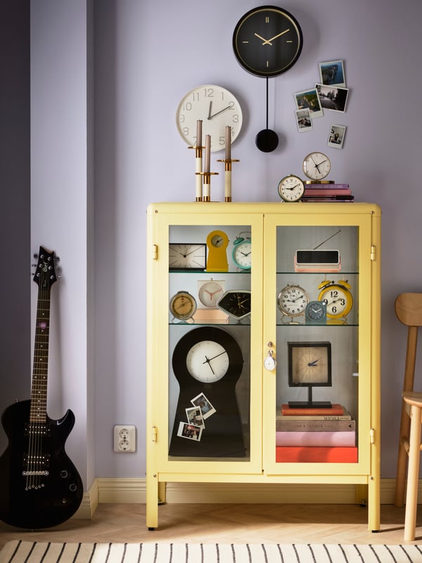 A living room with lilac-colored walls and a light yellow FABRIKÖR display cabinet with various clocks and alarm clocks inside.