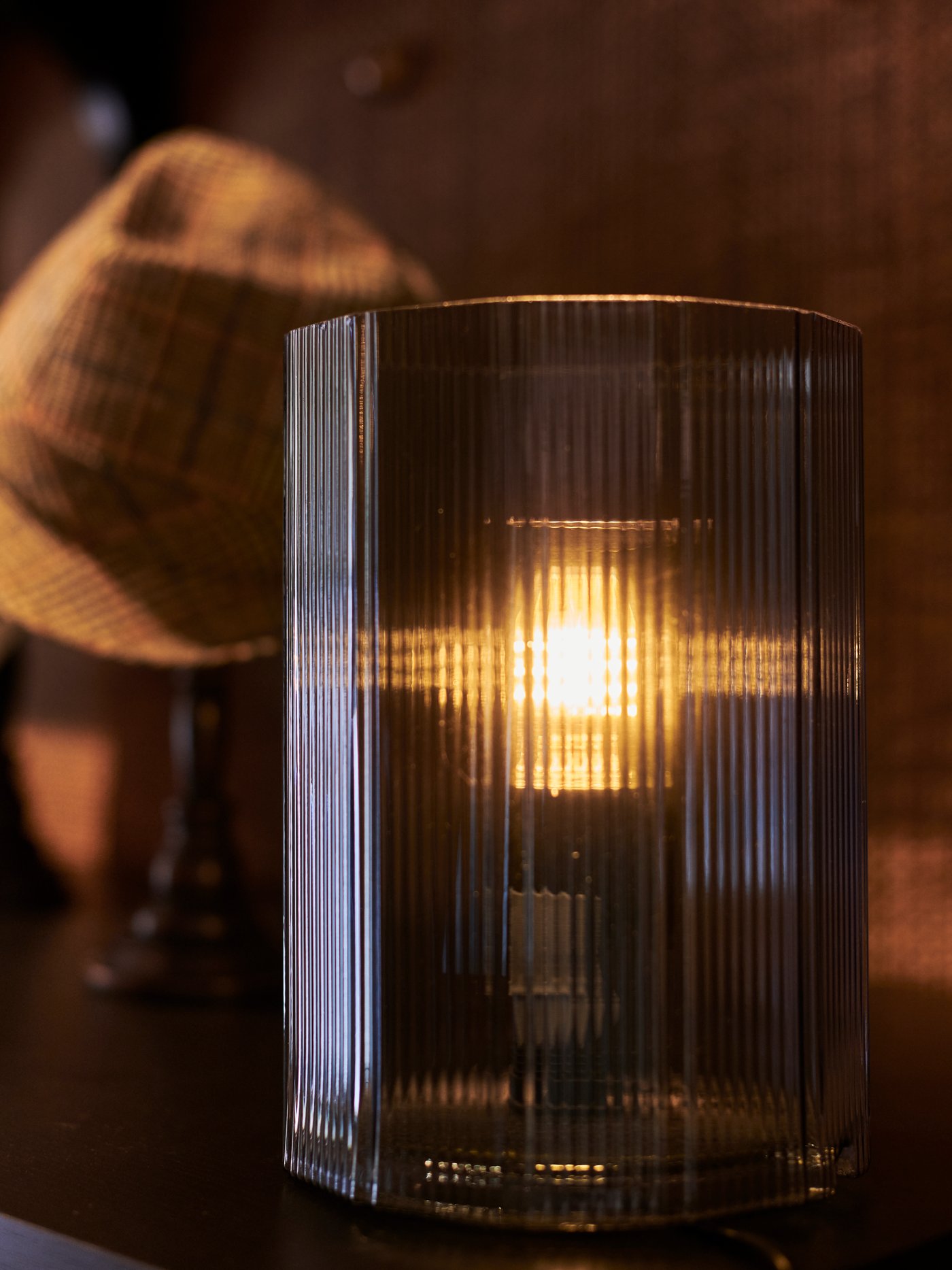 Modrá skleněná lampa MIKROKLIN na tmavém povrchu s hnědým kloboukem vzadu. Žárovka je rozsvícená a vydává jemné světlo.