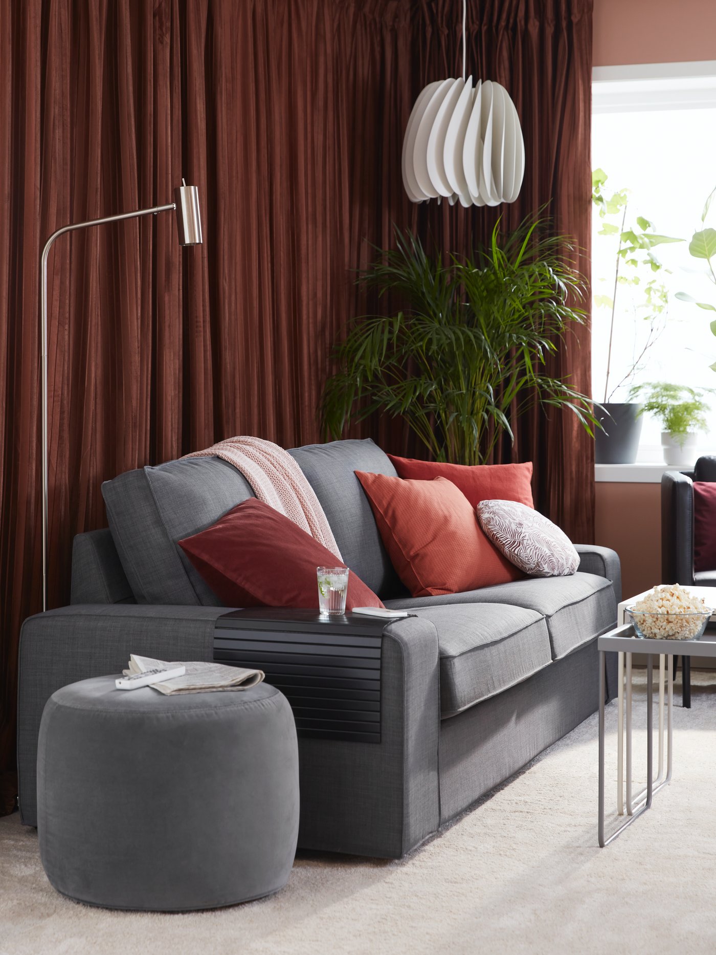 Mysig vardagsrumsmiljö med grå KIVIK soffa, färgglada kuddar i rött, rosa och vitt, grå fotpall, golvlampa och vit taklampa. Bruna gardiner och en grön krukväxt i bakgrunden.