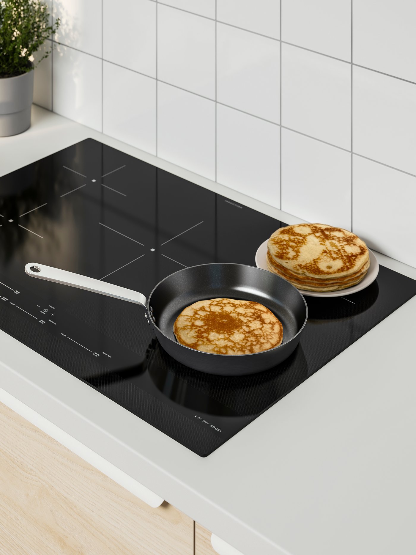 A pancake is being cooked in a frying pan on an induction cooktop. There is a plate with a stack of pancakes next to the pan.