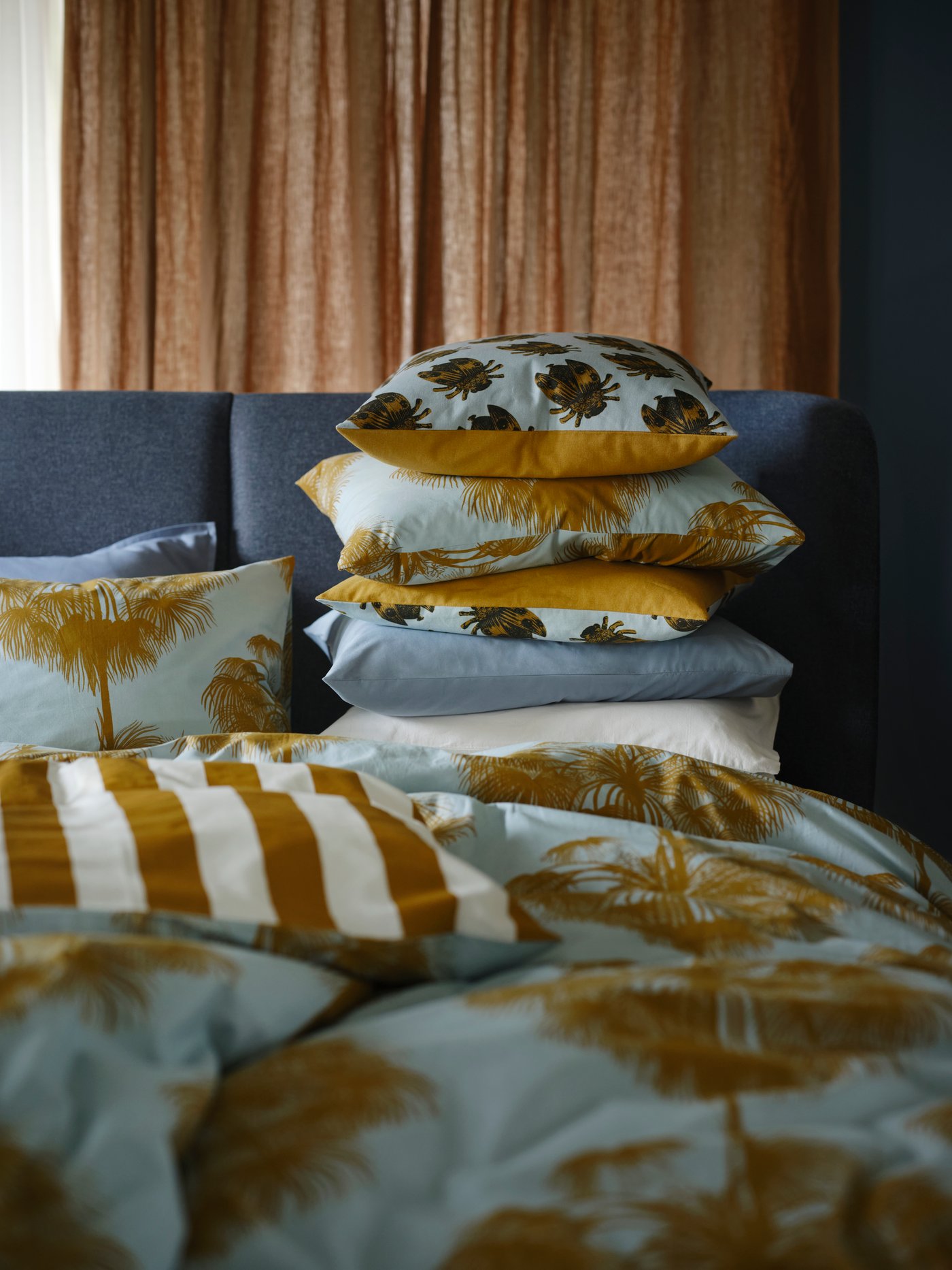 A blue bed that's made with ÄNGLATRUMPET bed linen, with pillows and cushions in NÄBBSPINNARE covers stacked in a pile.