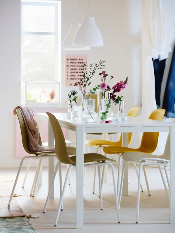 Una zona de comedor en tonos blancos con dos mesas MELLTORP blancas, lámparas de techo MELODI blancas y sillas LEIFARNE de diferentes colores.