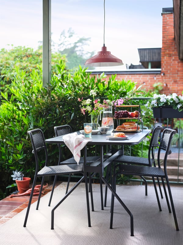 A VIHOLMEN table and four chairs on a terrace with a jug of water and a serving tray holding sandwiches and fruit on top.