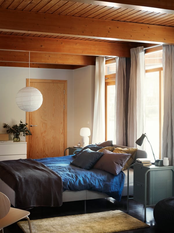 A bedroom with a light SLATTUM upholstered bed frame, blue bed linen, a throw, several pillows and a golden yellow rug.