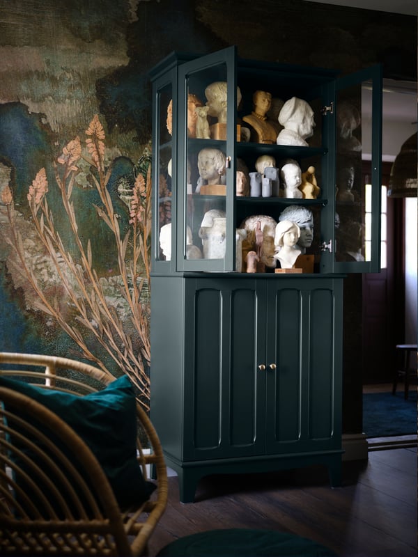 Une armoire vert foncé dont la partie supérieure possède des portes vitrées, dans laquelle différents bustes en terre glaise et en pierre sont conservés. 