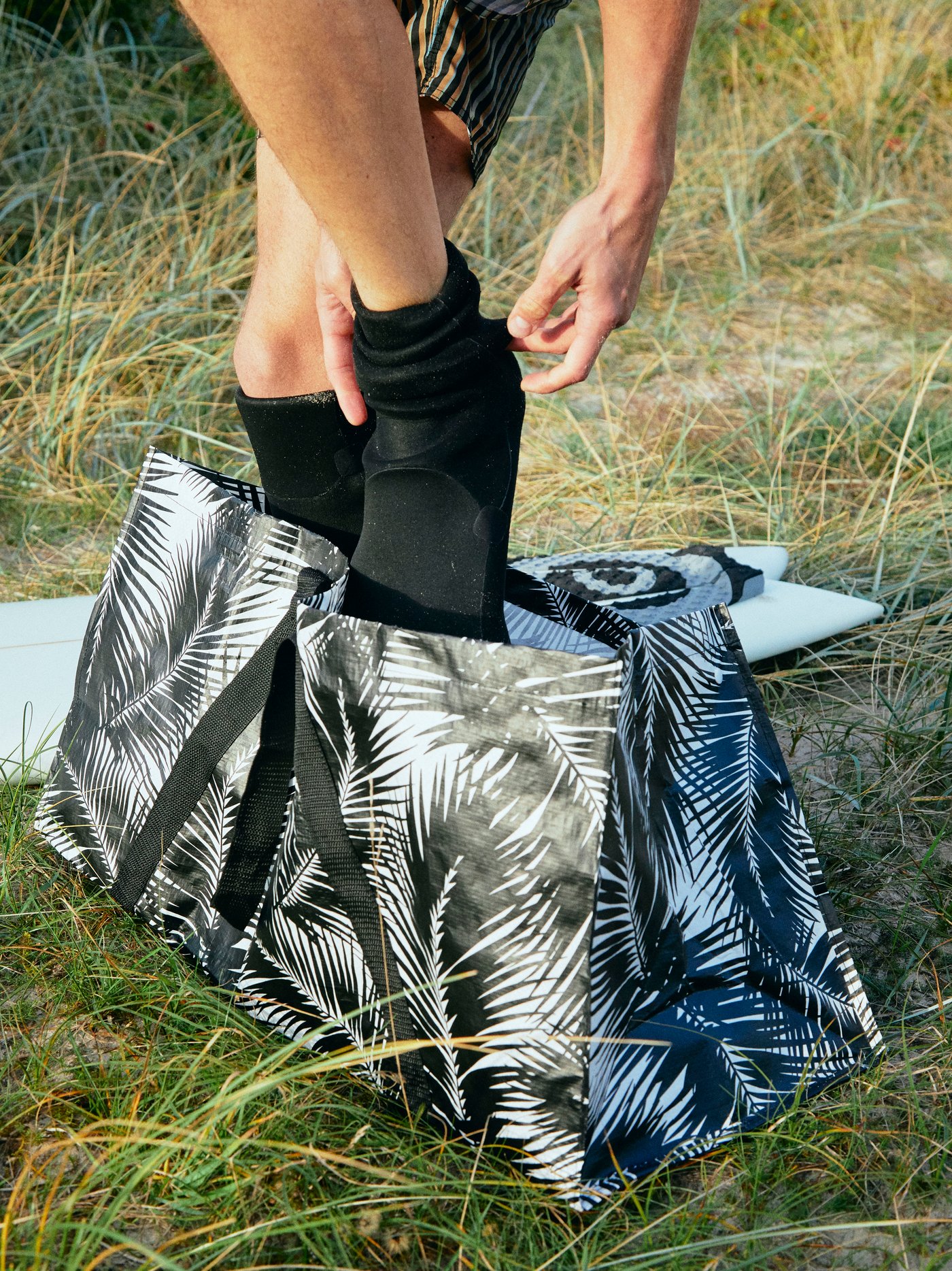 KÅSEBERGA bag in a black and white palm-inspired print placed on the grass with someone standing inside changing a wetsuit.