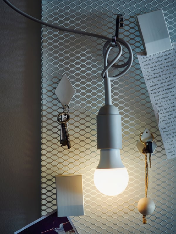 Lampadina a LED SOLHETTA accesa, appesa con un gancio a una rete metallica dove sono attaccati anche chiavi e promemoria.