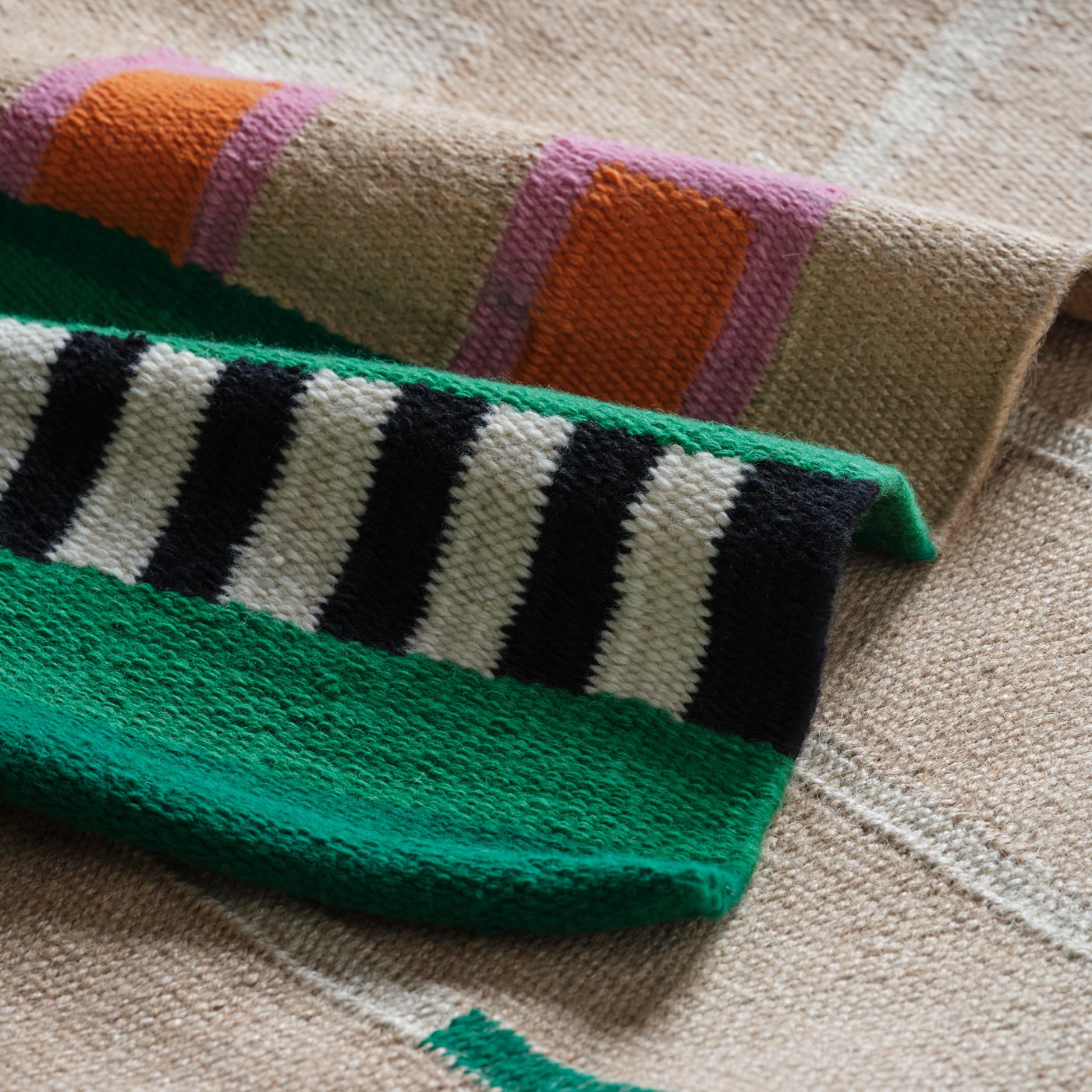 سجادة MÄVINN من الجوت غير مصبوغ وقطن بخطوط من الصوف الملون بالأسود والأبيض مع تفاصيل باللون الأخضر والوردي والبرتقالي.