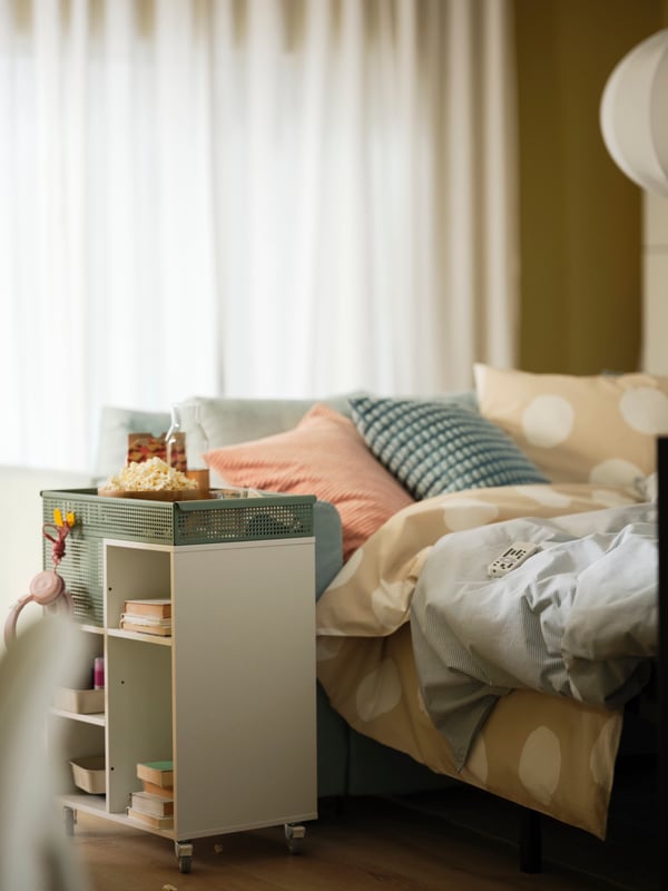 كنبة سرير 3 مقاعد VIMLE في وضع السرير عليه بياضات سرير مريحة. عربة ÖVNING بجانب الكنبة عليها الفشار والمشروبات.