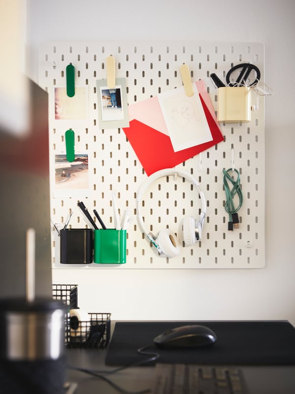 A white SKÅDIS pegboard is on a wall. Its accessories hold a headset, keys, scissors and more.