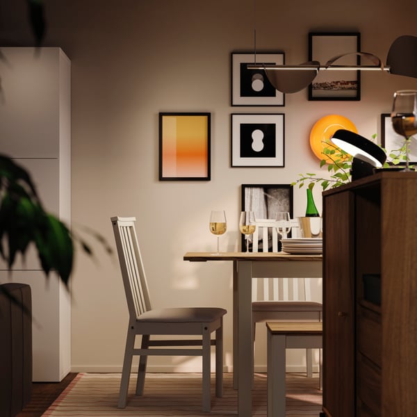 En spiseområde om aftenen med et ÅLHULT spisebordssæt med et bord med udtræk, en bænk og stole samt en rund, orange LED-lampe.