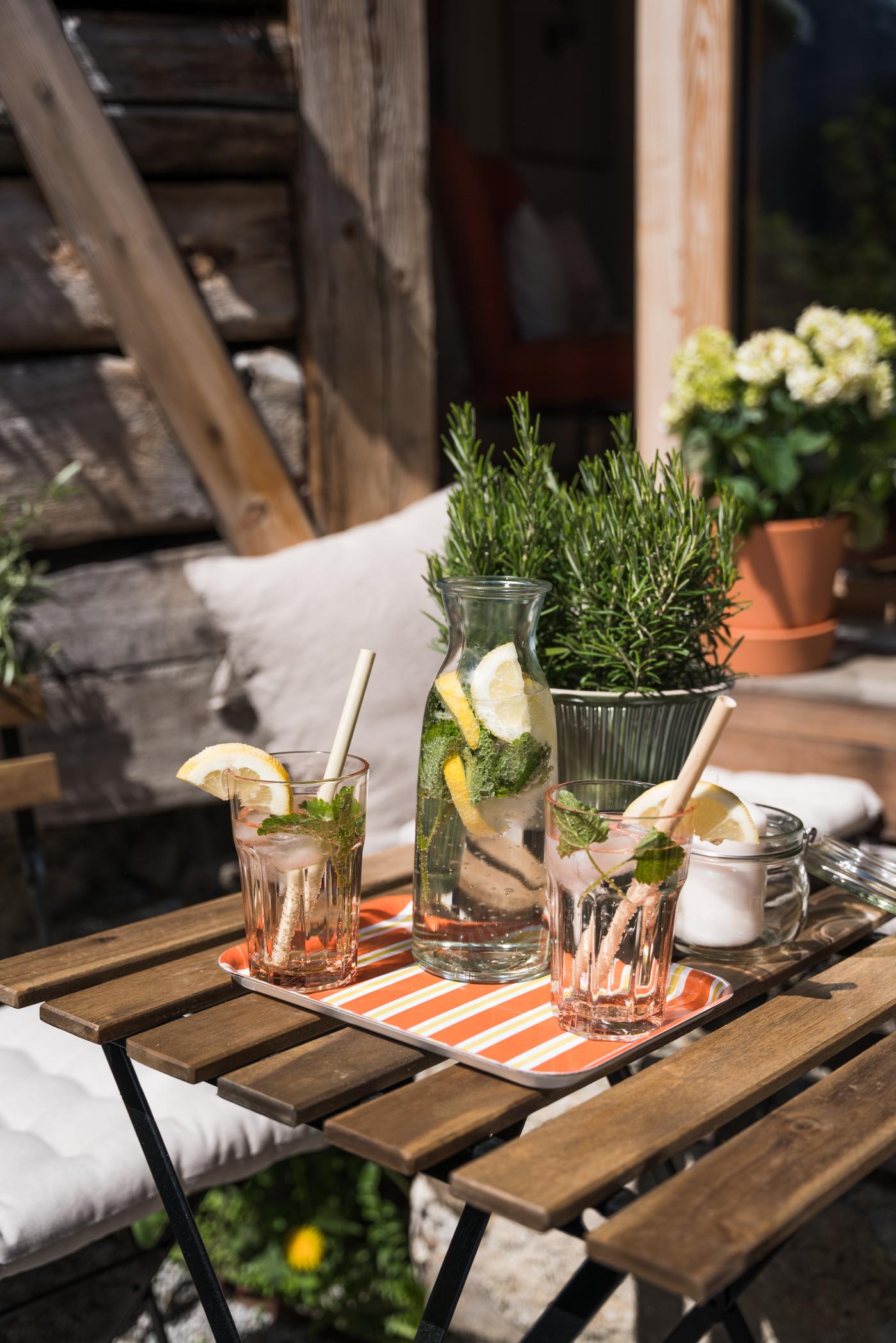 Erfrischende Getränke mit Zitrone und Minze auf einem Holztisch, neben Kräutern und Blumen im sonnigen Garten.