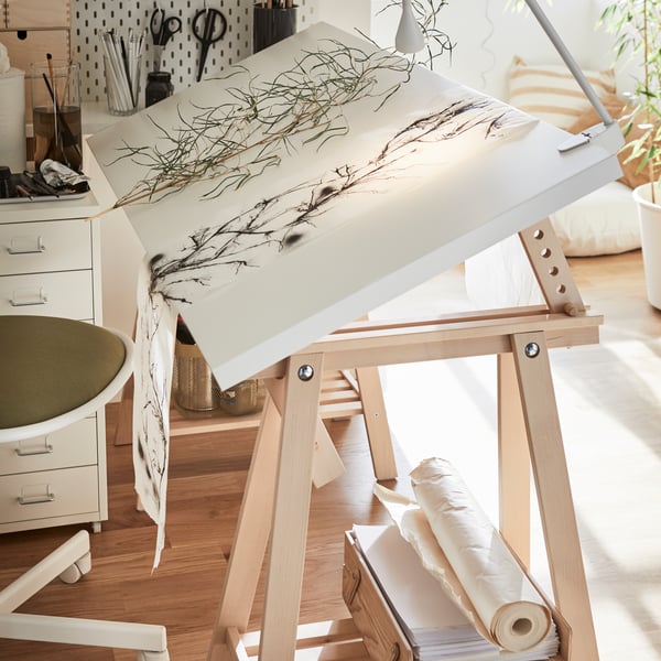 A white-and-birch LAGKAPTEN desk, its top tilted and with dried plants on paper on top and a white spotlight clamped to it.