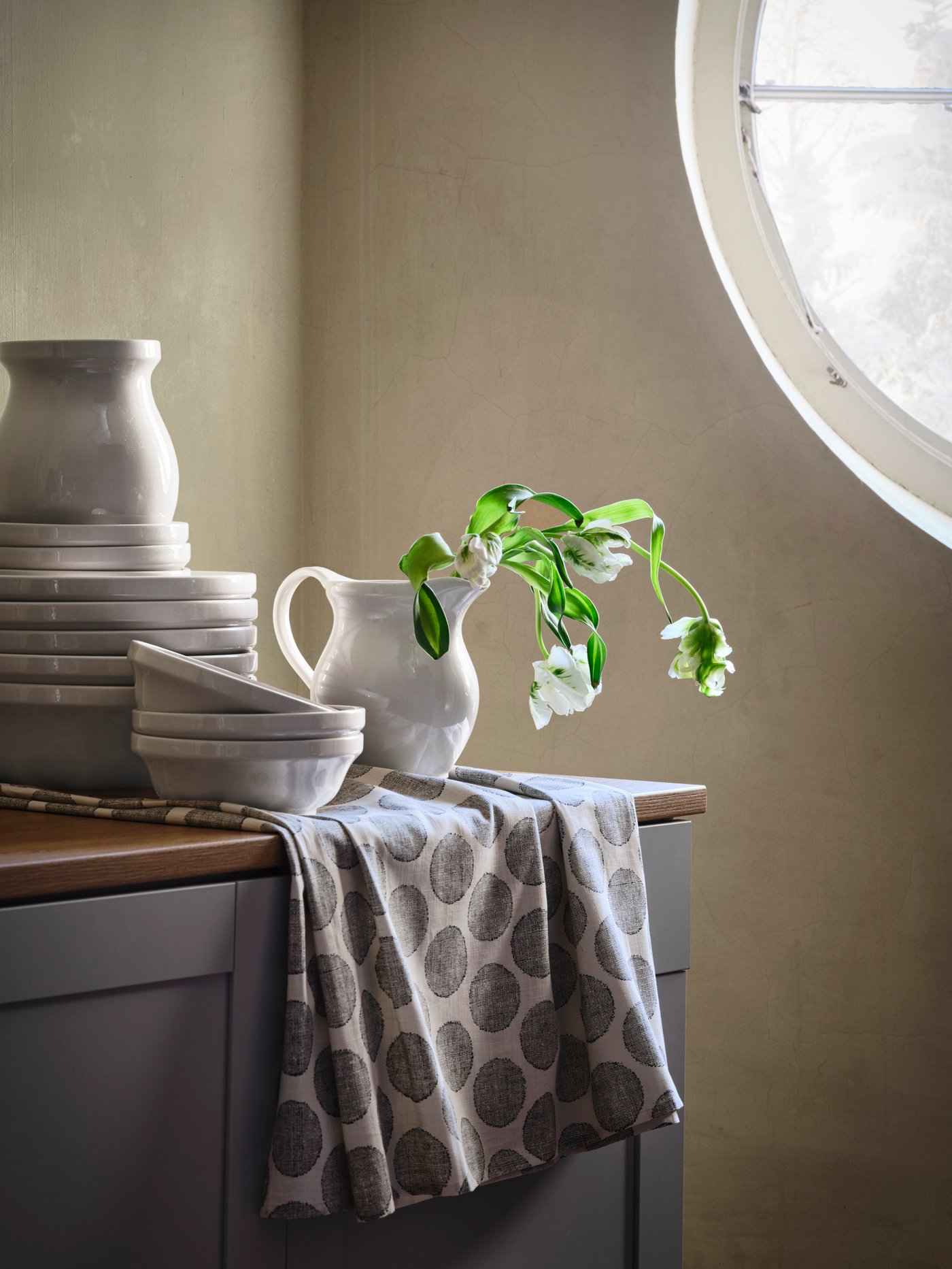 White DEKORERA bowls, vases and jugs are piled on top of each other on a kitchen sideboard, with flowers in one jug.